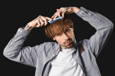 Genç dövmeli berber koyu arkaplanda saç tarıyor.