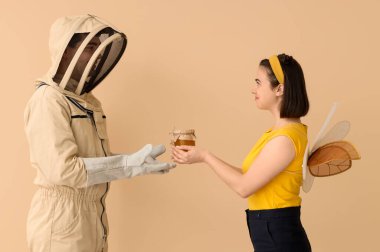 Male beekeeper with jar of honey and woman in bee costume on beige background clipart