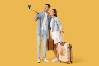 Güzel, genç, mutlu, bavullu turistler sarı arka planda selfie çekiyorlar.