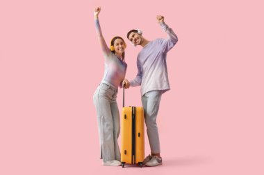 Güzel, genç, mutlu, kulaklıklı, bavullu, pembe arka planda dans eden iki turist.