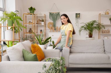 Genç, mutlu bir kadın, evdeki çiçeklerin yanındaki kanepede oturuyor.