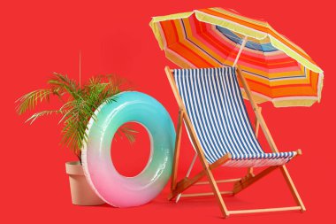 Güverte sandalyesi, plaj şemsiyesi, şişirilebilir yüzük ve kırmızı arka planda egzotik bitki.