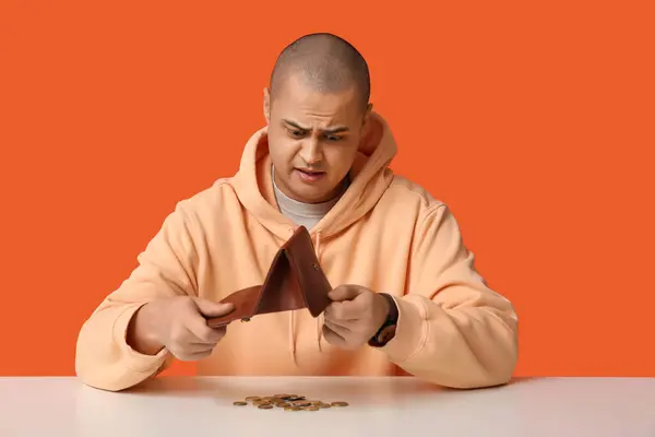 stock image Troubled young man emptying wallet with few coins on white table against orange background