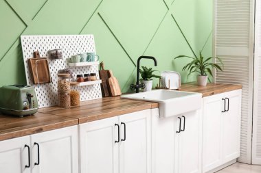 Mutfaktaki yeşil duvarın yanında tahta, yemek takımı ve ev bitkisi.