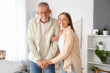 Kadın bakıcı huzurevinde yaşlı bir adama bastonla yardım ediyor.