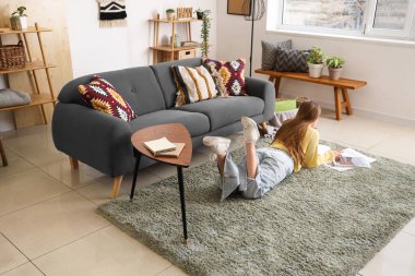 Genç bir kadın oturma odasında yerde uzanıyor ve dergileri okuyor.