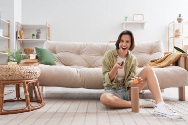 Genç bir kadın cep telefonu kullanıyor ve oturma odasında yerde oturuyor.