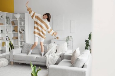 Genç, mutlu bir kadın, oturma odasında kanepede dans ediyor.