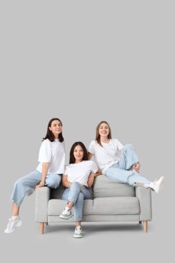 Şık kotlu genç kadınlar açık arka planda kanepede oturuyor.