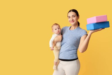 Yoga blokları olan genç ve güzel bir kadın küçük bebeğini sarı arka planda tutuyor.