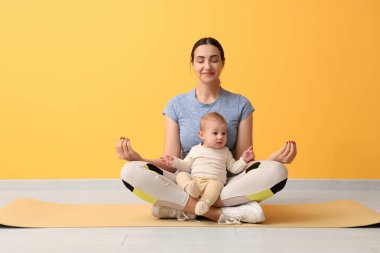 Güzel, genç, sportif bir kadın. Küçük bebeği, sarı duvarın yanında meditasyon yapıyor.