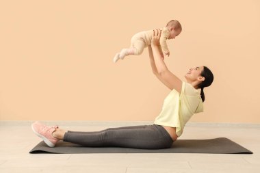 Bej duvarın yanında küçük bebeğiyle yoga yapan genç ve güzel bir kadın.