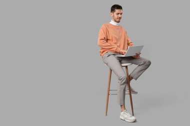 Gri arka planda dizüstü bilgisayarlı, şık şeftali rengi süveter giyen yakışıklı bir genç.