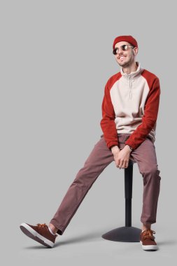 Güneş gözlüklü ve şapkalı yakışıklı, güler yüzlü genç adam gri arka planda oturuyordu.