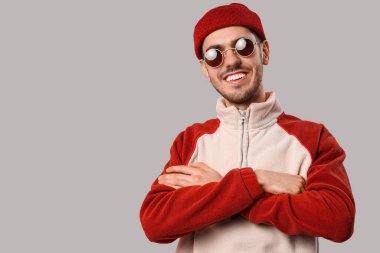 Yakışıklı, güler yüzlü, şık elbiseli, güneş gözlüklü ve gri arka planda poz veren genç bir adam.