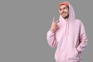 Gri arka planda zafer jesti gösteren zarif pembe kapüşonlu ve şeftali şapkalı yakışıklı genç adam.