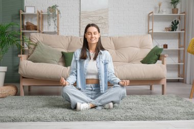 Genç bir kadın oturma odasındaki kanepenin yanında meditasyon yapıyor.