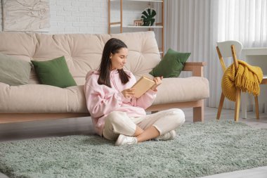 Oturma odasındaki kanepenin yanında kitap okuyan genç bir kadın.