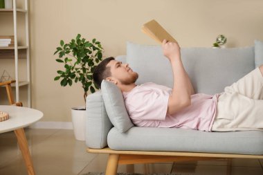 Genç adam kanepede uzanıp oturma odasında kitap okuyor.