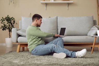 Genç adam kanepenin yanında oturuyor ve oturma odasında dizüstü bilgisayar kullanıyor.