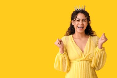 Sarı arka planda zarif balo elbisesi ve taç giyen mutlu Afro-Amerikan kadın.