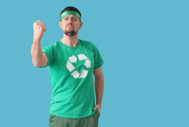 Tişörtlü, geri dönüşüm amblemi olan öfkeli genç adam mavi arka planda yumruk sallıyor. Ekoloji kavramı