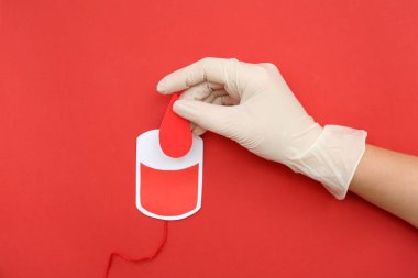 Kadın eli kauçuk eldivende, elinde kağıt damlası ve kırmızı arka planda kan nakli için kağıt kan torbası var. Dünya kan bağışı günü.
