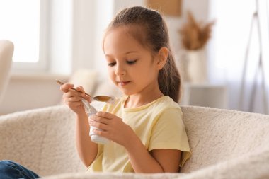 Cute little girl with spoon eating yogurt and sitting on sofa at home