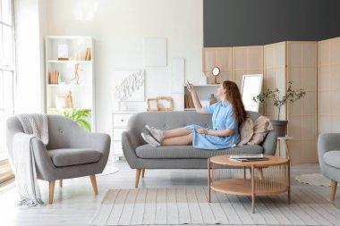 Evdeki kanepede oturan klimalı uzaktan kumandalı genç bir kadın.