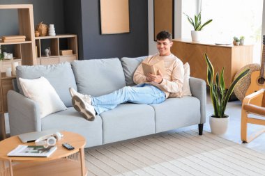 Genç adam evdeki kanepede kitap okuyor.