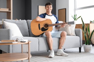 Genç adam evdeki kanepede gitar çalıyor.