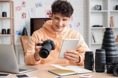 Modern kamera, tablet ve dizüstü bilgisayarlı profesyonel mutlu erkek fotoğrafçı stüdyoda oturuyor.