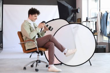 Modern kameralı profesyonel mutlu erkek fotoğrafçı ve stüdyoda sandalyede oturan yumuşak kutular.
