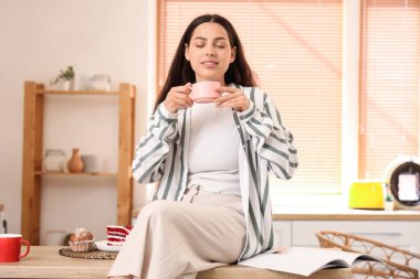 Genç, mutlu bir kadın, elinde bir fincan taze kahve ve mutfakta tatlı okuyarak.