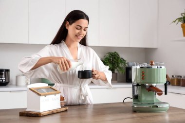 Modern bir makineyle genç ve güzel bir kadın mutfakta bir fincan taze kahveye süt dolduruyor.