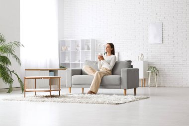 Genç ve güzel bir kadın evdeki rahat koltukta kahve içiyor.