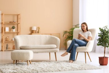 Genç ve güzel bir kadın cep telefonu kullanıyor ve oturma odasında koltukta oturuyor.