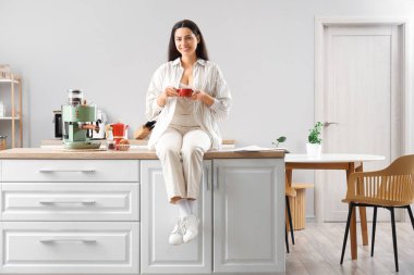 Elinde bir fincan sıcak espresso ve kahve makinesiyle evdeki mutfak tezgahında oturan genç ve güzel bir kadın.