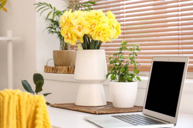 Modern dizüstü bilgisayarı ve beyaz masadaki sarı narsisli çiçekleri olan rahat çalışma alanı.