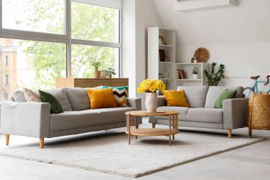 Aydınlık oturma odasının içinde gri kanepeler ve ahşap sehpanın üzerinde sarı narsisli çiçekler.