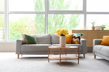 Aydınlık oturma odasının içinde gri kanepeler ve ahşap sehpanın üzerinde sarı narsisli çiçekler.