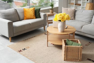 Oturma odasının içinde gri kanepeler, dizüstü bilgisayarlar ve ahşap sehpada sarı narsisli çiçekler.