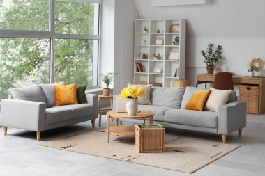 Oturma odasının içinde gri kanepeler, dizüstü bilgisayarlar ve ahşap sehpada sarı narsisli çiçekler.