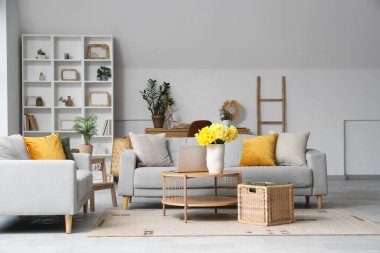 Oturma odasının içinde gri kanepeler, dizüstü bilgisayarlar ve ahşap sehpada sarı narsisli çiçekler.