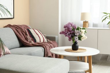 Kahve masasının üzerinde bir buket leylak dalları, rahat bir kanepe ve yastıklar olan güzel bir oturma odası.
