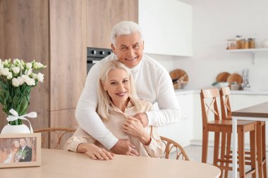 Olgun bir adam, evlilik yıldönümlerinde mutfakta karısına sarılıyor.