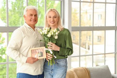 Fotoğraf çerçeveli ve çiçekli olgun bir çift evde evlilik yıldönümlerini kutluyor.