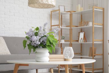 Güzel bir çiçek buketi, kitaplar, masa üzerinde lamba, kanepe ve oturma odasının içinde raf ünitesi olan beyaz vazo.