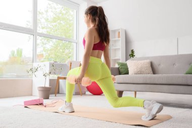 Dambılı evde ve arka planda antrenman yapan genç bir bayan.