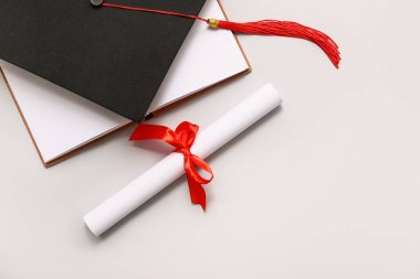 Kırmızı kurdeleli diploma, açık kitap ve beyaz arka planda mezuniyet şapkası.